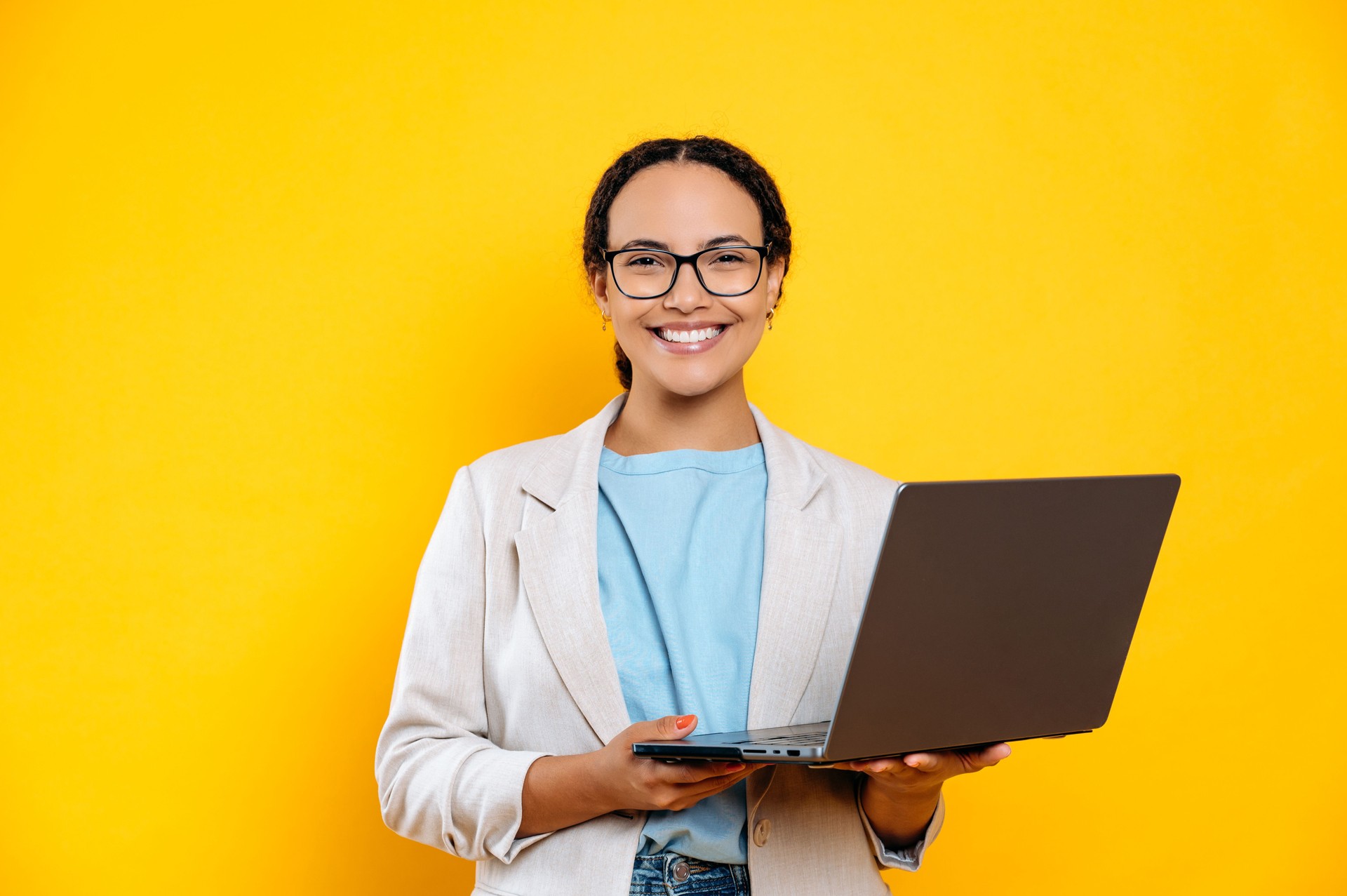 Lovely confident successful mixed race brunette woman, business lady, programmer, creative manager, with glasses, hold in hands open laptop, stand on isolated yellow background, looks at camera, smile