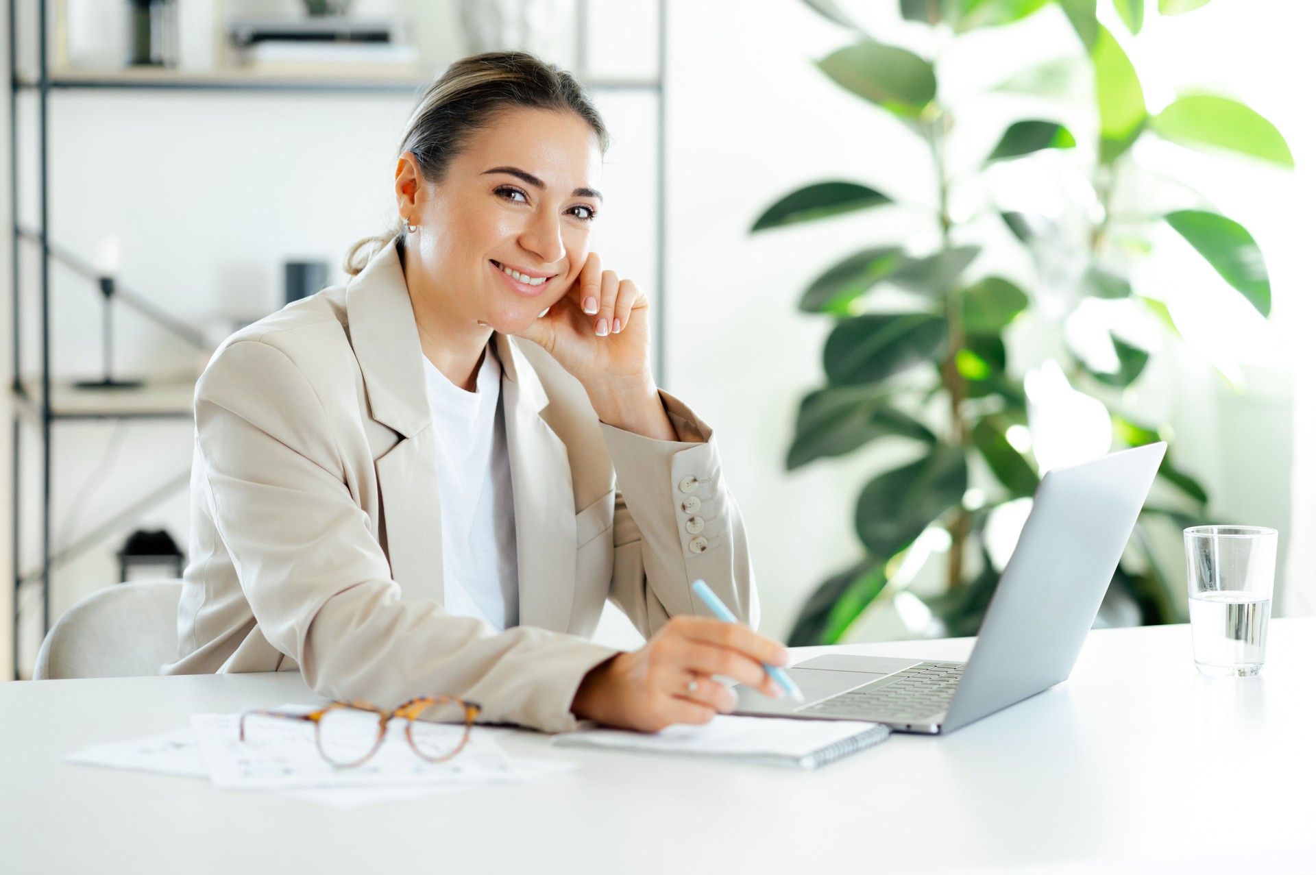 femme caucasienne professionnelle positive dans un costume, femme d’affaires, travaillant sur un ordinateur portable tout en étant assise à un bureau moderne dans un bureau moderne lumineux, regardant la caméra et souriant amicalement