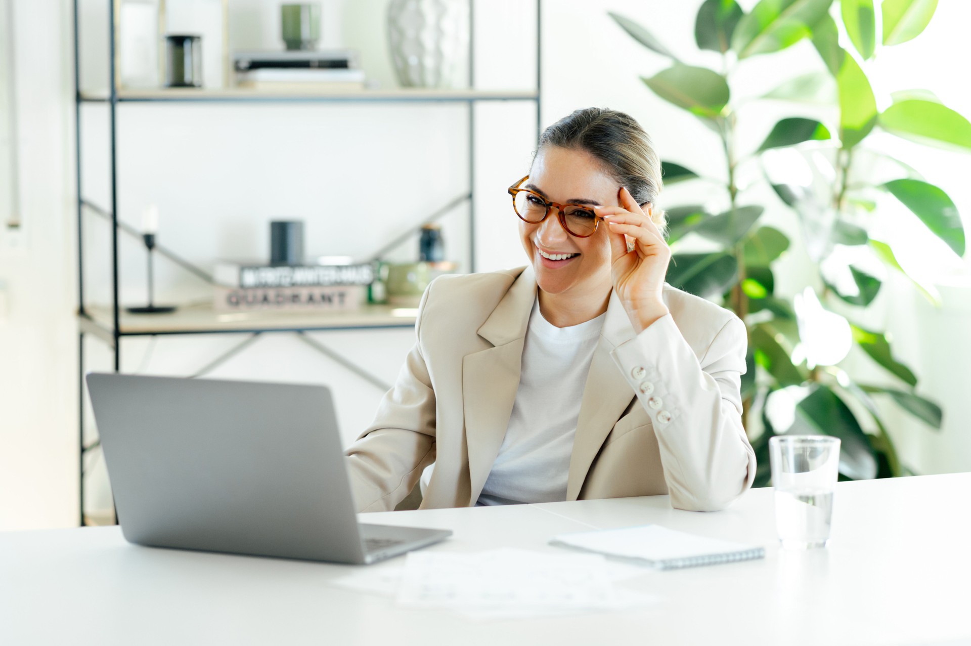 Jolie femme d’affaires caucasienne joyeuse et réussie dans un costume élégant et des lunettes, assise à son lieu de travail avec un ordinateur portable dans un bureau moderne, regarde l’écran, souriant joyeusement